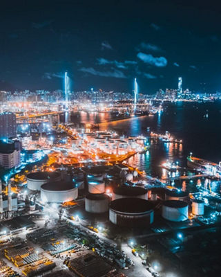 Nighttime view of an industrial port city with illuminated ships, tanks, and skyscrapers, representing global industry and progress.