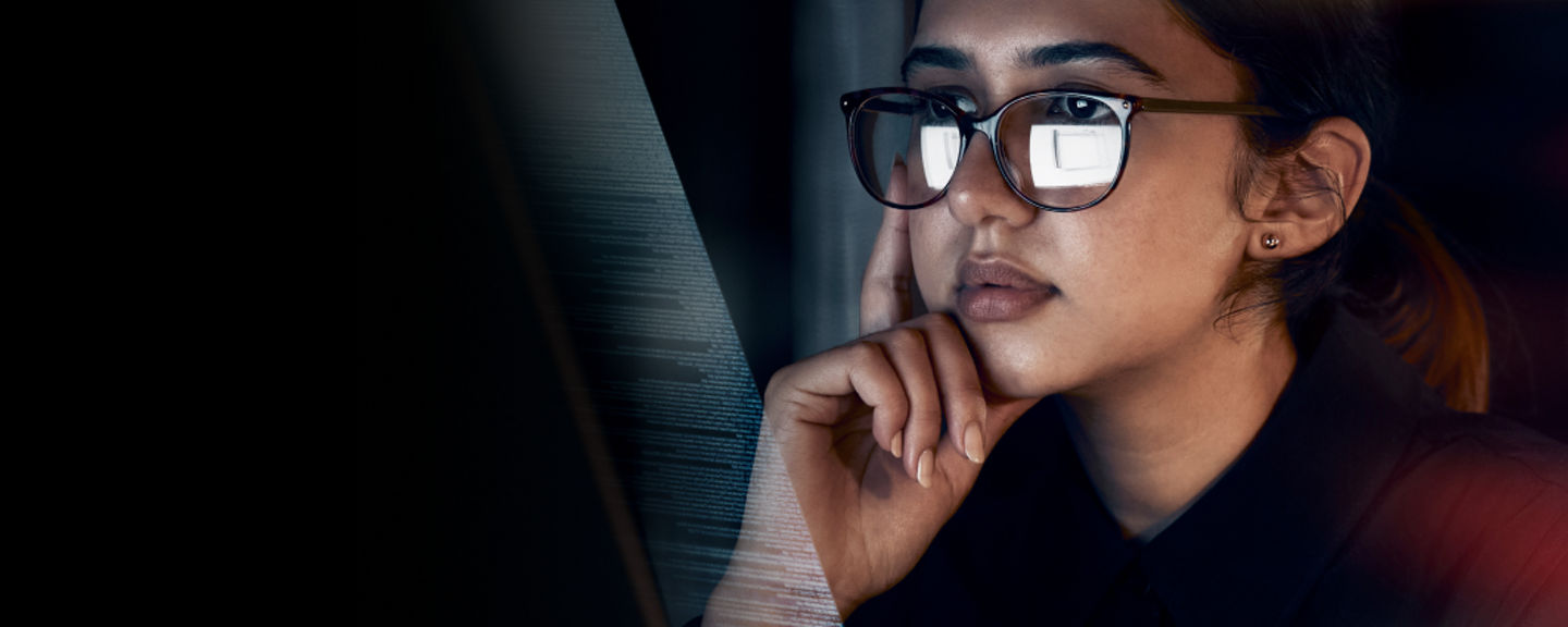 Focused professional woman wearing glasses, analyzing information on a computer screen in a dimly lit setting.