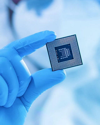 Gloved hand holding a computer microchip in a cleanroom environment, representing semiconductor technology.