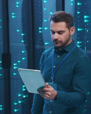 IT professional standing in a data center with illuminated server racks, holding a tablet and analyzing system information.