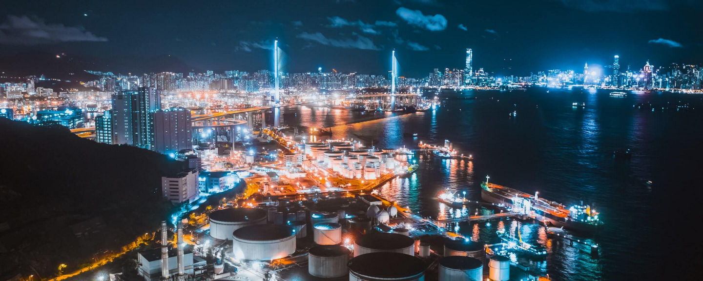 Nighttime view of an industrial port city with illuminated ships, tanks, and skyscrapers, representing global industry and progress.