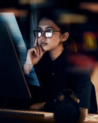 Focused professional woman wearing glasses, analyzing information on a computer screen in a dimly lit setting.