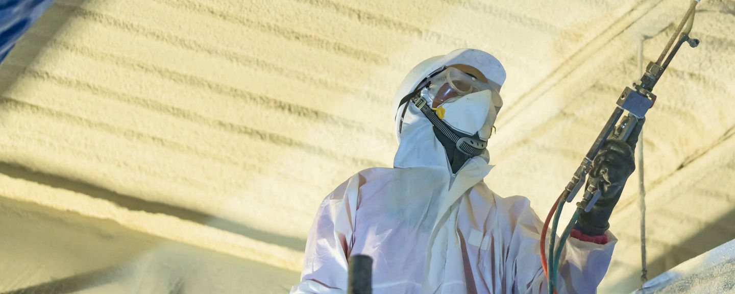 Worker in protective gear spraying insulation foam on a ceiling inside a construction site, representing energy-efficient building insulation.