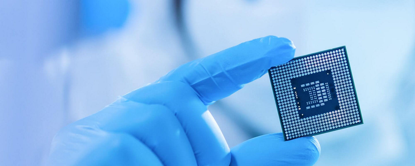 Gloved hand holding a computer microchip in a cleanroom environment, representing semiconductor technology.
