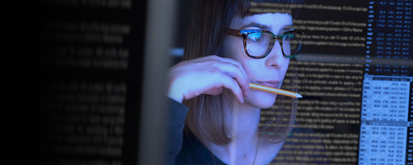 Focused professional woman wearing glasses working on a computer surrounded by lines of code, representing digital talent and career opportunities in technology.