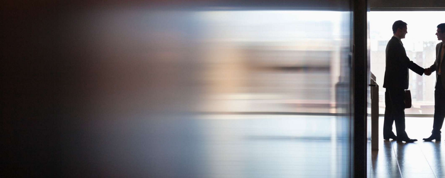 Two business professionals shaking hands in an office hallway with city buildings visible through large windows, representing partnership.