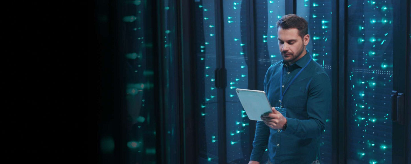 IT professional standing in a data center with illuminated server racks, holding a tablet and analyzing system information.